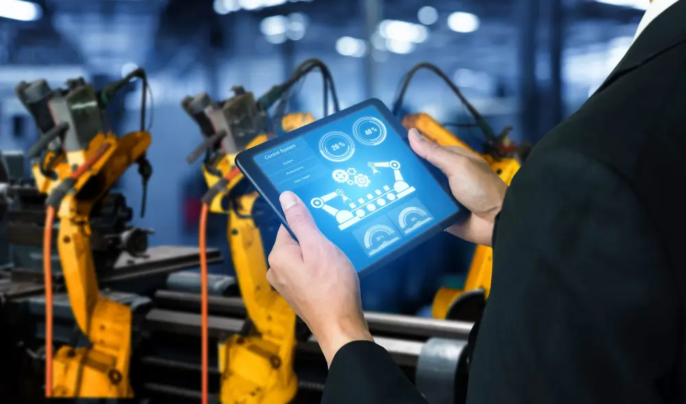Profissional de terno segura tablet exibindo painel de controle industrial, com braços robóticos amarelos ao fundo em ambiente de fábrica.