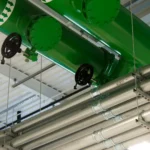 Tubulação verde de grande diâmetro com válvulas de volante e eletrodutos metálicos correndo no teto de uma instalação industrial