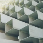 Vista em ângulo de fachada de edifício com painéis triangulares metálicos formando padrão repetitivo, luz solar incidindo pela lateral